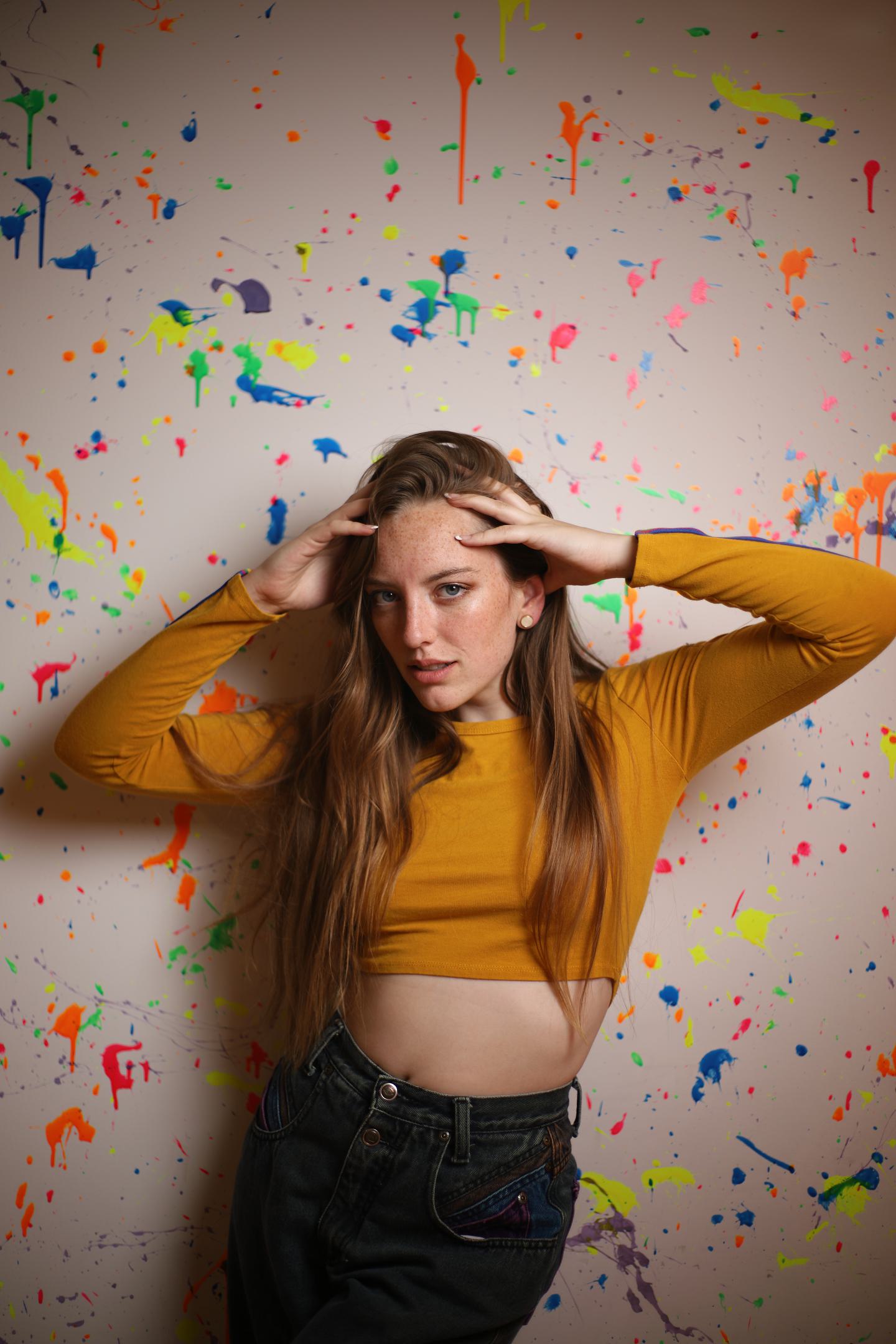 Bold fashion portrait of woman in mustard yellow crop top hands on head direct gaze against paint splatter wall indoor studio Grand Rapids Michigan