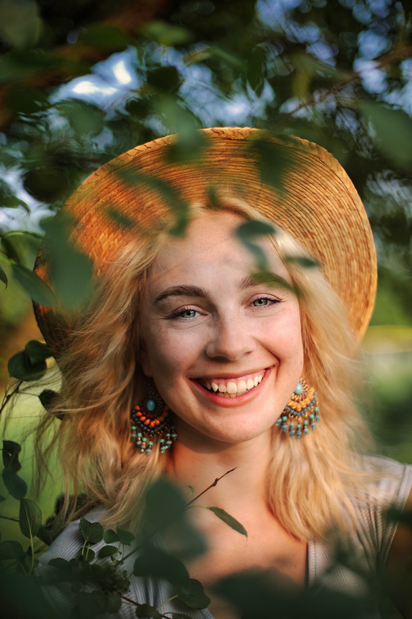 Joyful golden hour fashion portrait of woman in straw hat smiling through green leaves at Rotary Park Livonia Michigan