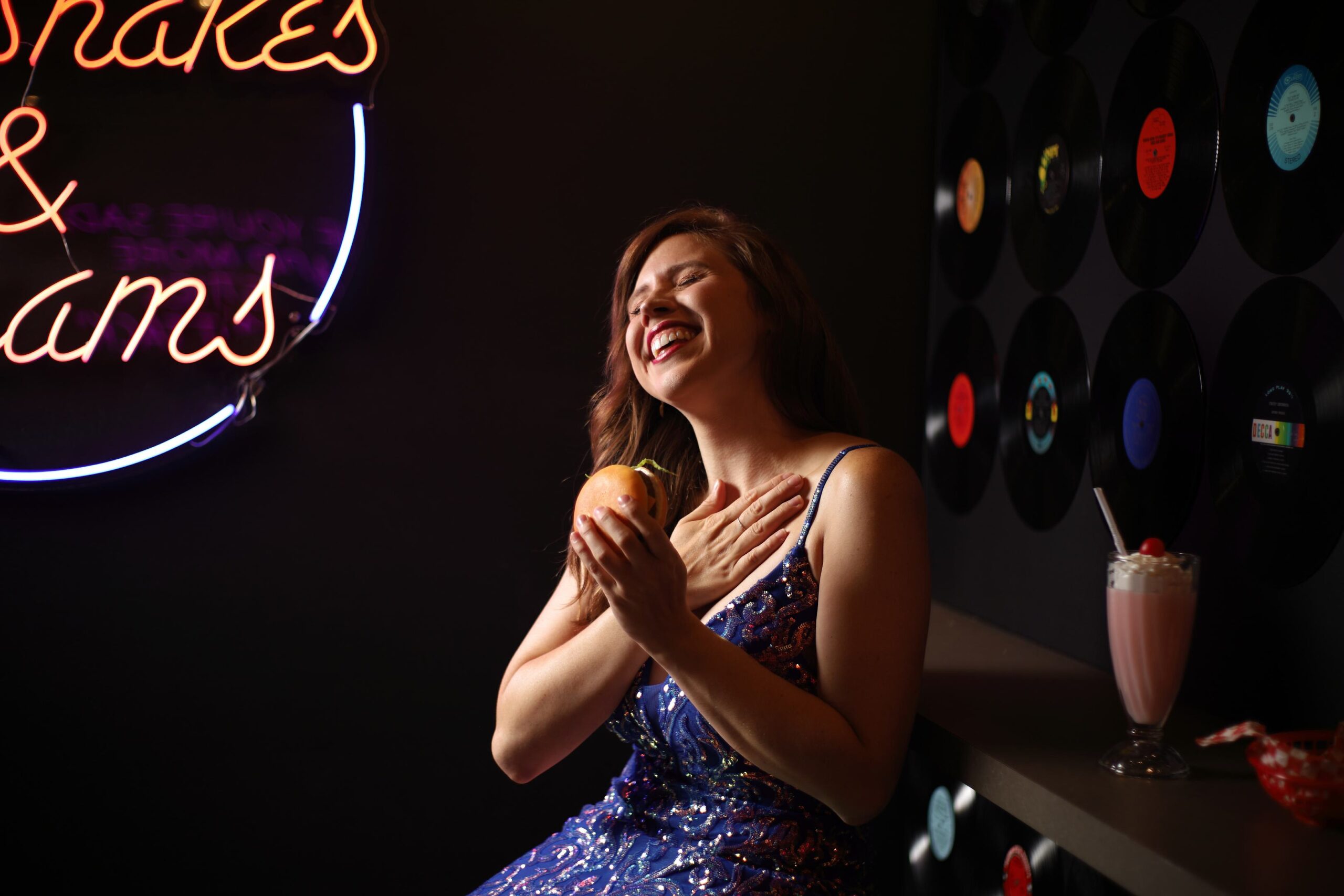 Candid fashion portrait of woman in blue sequin gown laughing eyes closed holding burger to chest at retro diner Grand Rapids Michigan