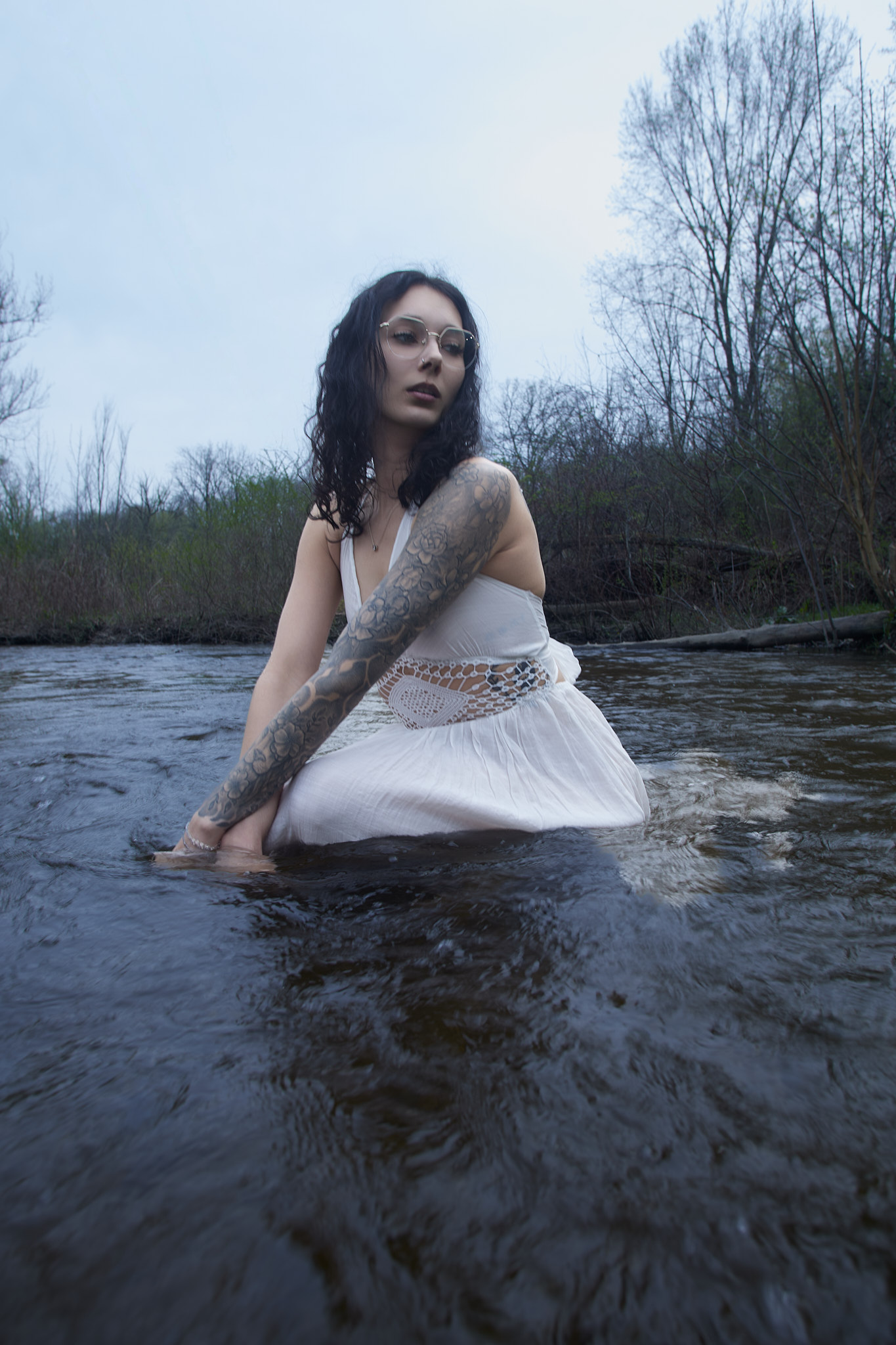 Moody outdoor fashion portrait woman soaking white boho dress rose tattoo sleeve crouching river downward gaze Detroit Metro Michigan