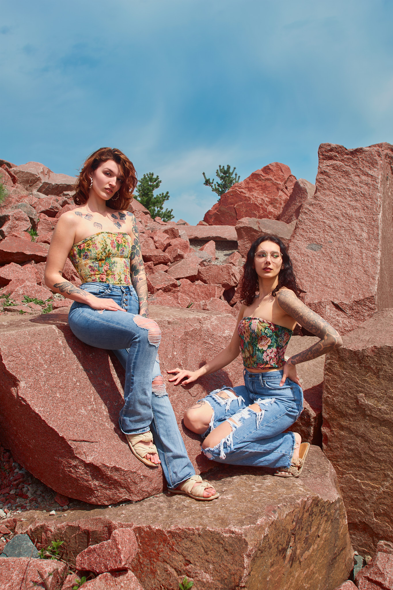 dynamic editorial fashion portrait two tattooed models staggered heights red granite quarry auburn hair yellow floral corset distressed flare jeans leg raised dark hair glasses jewel-toned floral brocade corset wide-leg distressed jeans hand on rock direct gaze Detroit Metro Michigan