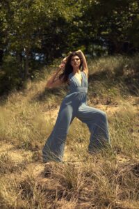 Full body fashion portrait of woman in blue wide-leg jumpsuit arms raised among dune grass at Hoffmaster State Park west Michigan