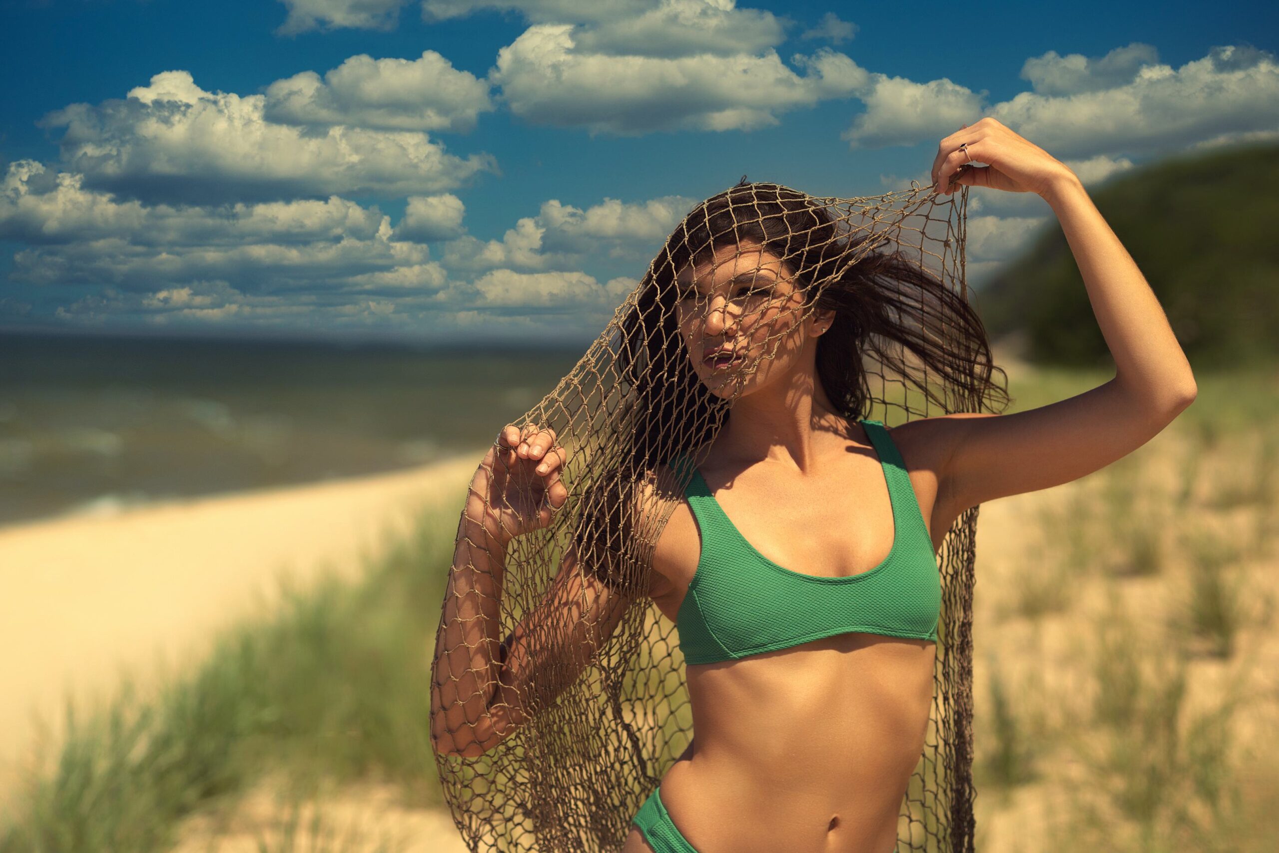 Creative fashion portrait of woman in green bikini holding fishing net over face on west Michigan sand dunes