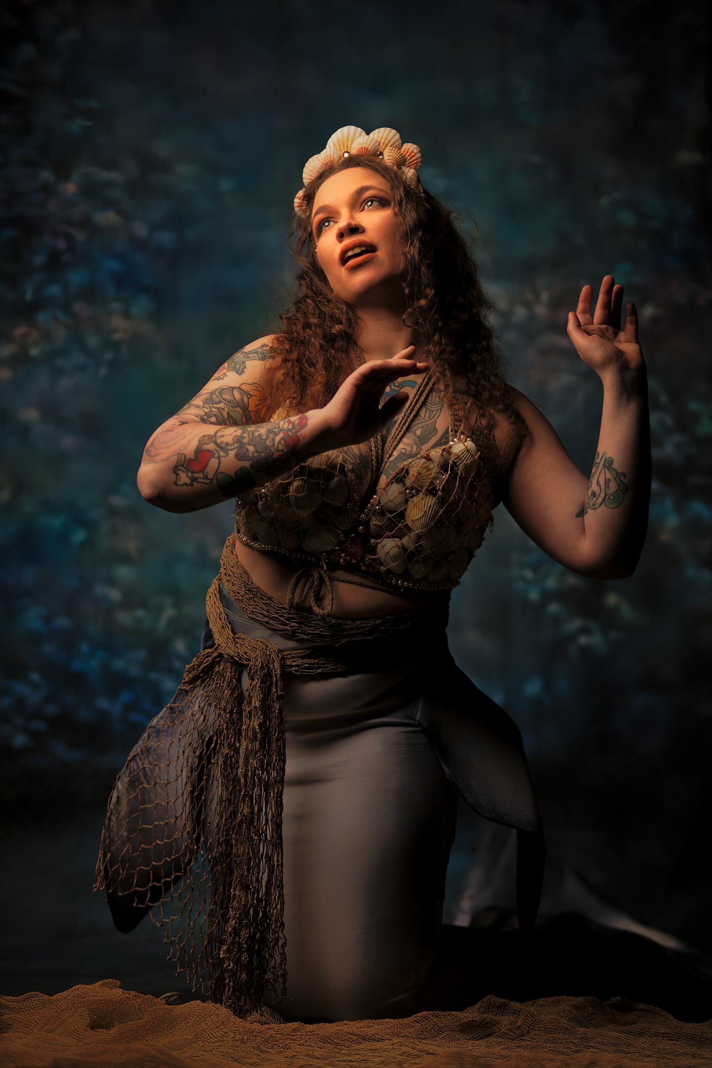 Creative mermaid concept fashion portrait of tattooed woman in shell crown and seashell top kneeling on sand against blue backdrop Oakland County Michigan