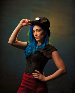 Bold steampunk concept fashion portrait of woman with blue hair tipping black top hat with goggles direct gaze Oakland County Michigan