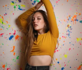Indoor Creative Fashion Photography — Grand Rapids Michigan Paint Wall Full body fashion portrait of woman in mustard yellow crop top arm raised against colorful paint splatter wall indoor studio Grand Rapids Michigan