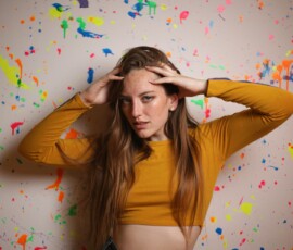 Bold Indoor Fashion Portrait — Grand Rapids Michigan Paint Splatter Wall Bold fashion portrait of woman in mustard yellow crop top hands on head direct gaze against paint splatter wall indoor studio Grand Rapids Michigan