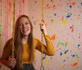Joyful Indoor Swing Fashion Portrait — Grand Rapids Michigan Joyful fashion portrait of woman in mustard yellow crop top laughing on yellow chain swing against paint splatter wall indoor studio Grand Rapids Michigan