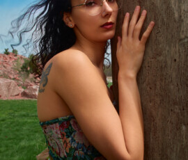 Detroit Metro Michigan Moody Outdoor Fashion Photography — Dark Floral Brocade Corset Solo Over-Shoulder Look moody solo outdoor fashion portrait dark-haired tattooed model wire-frame glasses red lip structured jewel-toned floral brocade corset high-waist distressed jeans back to camera hand pressed weathered tree trunk over-shoulder gaze butterfly shoulder tattoo blue sky Detroit Metro Michigan