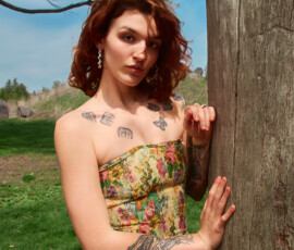 Detroit Metro Michigan Editorial Outdoor Fashion Photography — Yellow Floral Corset Solo Tattooed Model Tree close editorial outdoor fashion portrait auburn-haired tattooed model yellow floral corset high-waist distressed flare jeans leaning weathered tree trunk hand on bark ornate drop earrings botanical butterfly chest tattoos direct gaze blue sky Detroit Metro Michigan