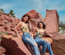 Detroit Metro Michigan Editorial Rock Quarry Fashion Photography — Duo Floral Corset High-Waist Denim Look editorial outdoor fashion portrait two tattooed models floral corset tops distressed blue jeans seated upright red granite quarry auburn hair ornate earrings arm tattoos dark hair wire-frame glasses delicate necklace direct gaze blue sky Detroit Metro Michigan