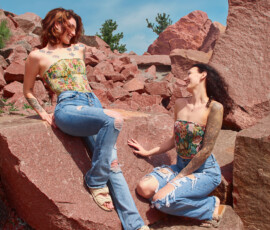 Detroit Metro Michigan Candid Editorial Fashion Photography — Duo Floral Corset Denim Laughing Natural Moment candid editorial fashion portrait two tattooed models floral corset tops distressed blue jeans laughing red granite quarry auburn hair elevated smiling dark hair glasses below laughing natural chemistry blue sky Detroit Metro Michigan