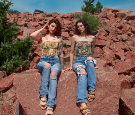 Detroit Metro Michigan Editorial Outdoor Fashion Photography — Duo Side by Side Flat Rock Floral Corset Denim wide editorial fashion portrait two tattooed models seated side by side flat red granite rock legs relaxed auburn hair hand raised ear dark hair wire-frame glasses floral corset tops distressed jeans red rock quarry green pines Detroit Metro Michigan