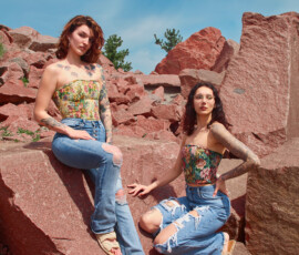 Detroit Metro Michigan Editorial Fashion Photography — Staggered Rock Duo Floral Corset Distressed Denim dynamic editorial fashion portrait two tattooed models staggered heights red granite quarry auburn hair yellow floral corset distressed flare jeans leg raised dark hair glasses jewel-toned floral brocade corset wide-leg distressed jeans hand on rock direct gaze Detroit Metro Michigan