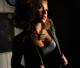 Traverse City Michigan Moody Indoor Fashion Photography Moody fashion portrait of woman with curly hair in black leather jacket and bodysuit with gold chain necklace world clock wall background Traverse City Michigan