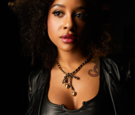 Traverse City Michigan Close Up Fashion Portrait — Gold Heart Necklace Striking close up fashion portrait of woman with curly hair in black leather jacket and gold heart charm necklace direct gaze black background Traverse City Michigan