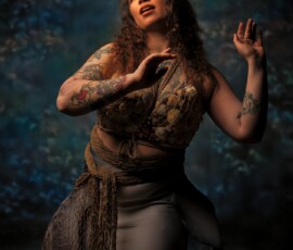 Oakland County Michigan Creative Concept Fashion Photography — Mermaid Session Creative mermaid concept fashion portrait of tattooed woman in shell crown and seashell top kneeling on sand against blue backdrop Oakland County Michigan