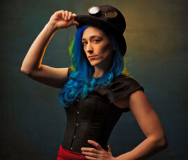 Bold Steampunk Studio Fashion Portrait — Oakland County Michigan Bold steampunk concept fashion portrait of woman with blue hair tipping black top hat with goggles direct gaze Oakland County Michigan