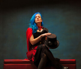 Oakland County Michigan Steampunk Concept Studio Photography — Holding Hat Contemplative steampunk concept fashion portrait of woman with blue hair seated on red ottoman holding black top hat with goggles Oakland County Michigan